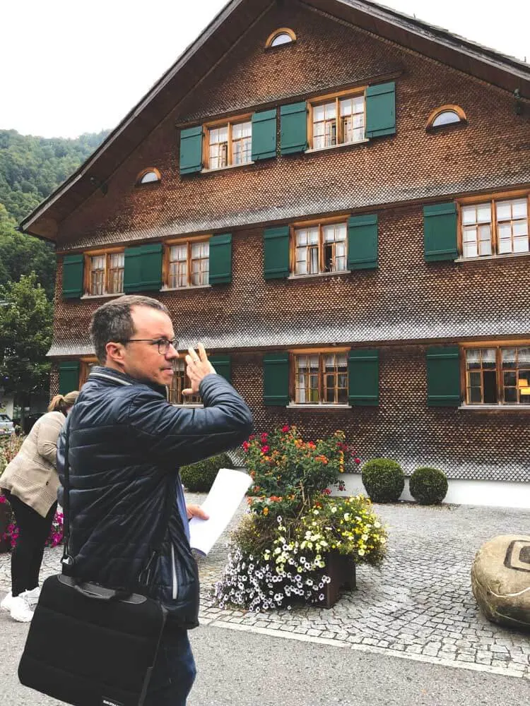 A man pointing to a wooden house on a guided tour