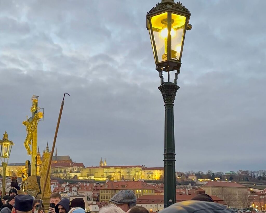 A man in a vintage uniform lights a gas streetlamp in Prague using a long pole, while a small crowd watches during the evening lamp-lighting ceremony.