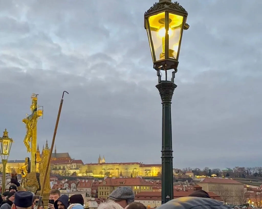 A man in a vintage uniform lights a gas streetlamp in Prague using a long pole, while a small crowd watches during the evening lamp-lighting ceremony.