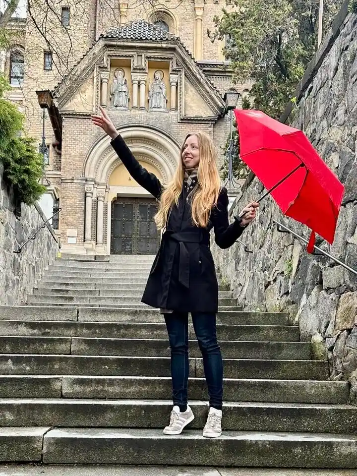 A woman in a black coat stands on a stone staircase, holding a bright red umbrella and joyfully gesturing toward the sky. Behind her, a historic brick church with arched windows and statues adds to the old-world charm of Prague in spring.