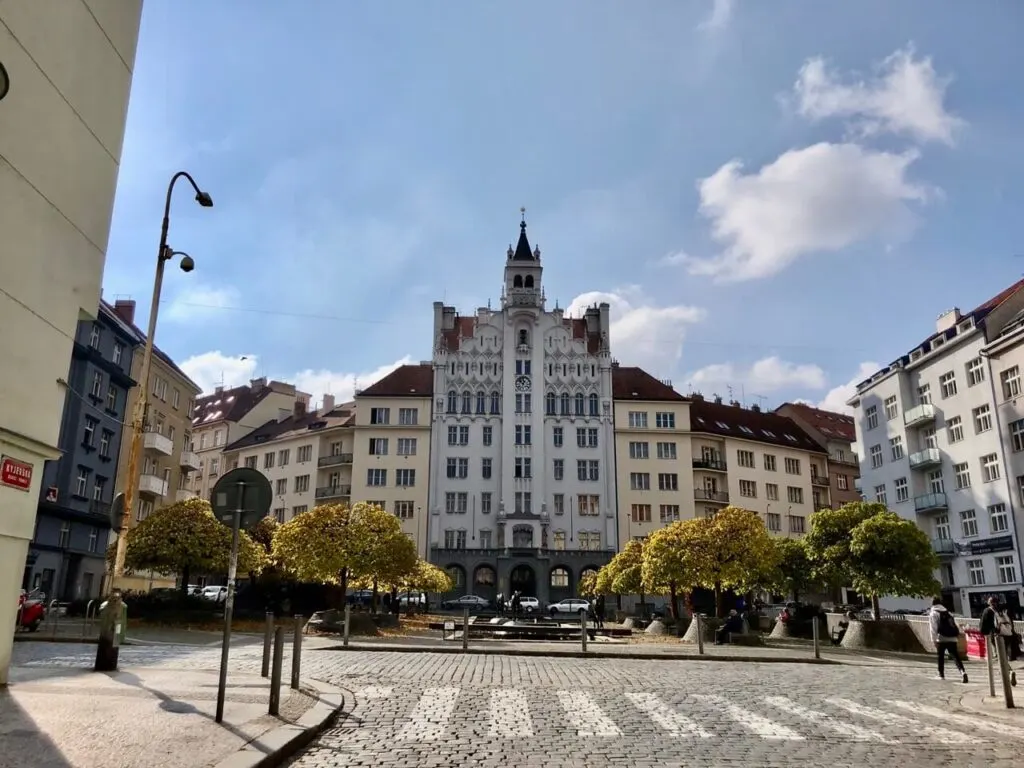 Cobblestone plaza in Prague with neatly trimmed trees and a grand white building featuring Gothic Revival architecture under a blue sky.