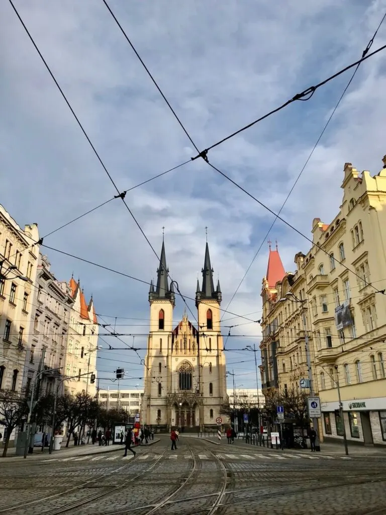 Gothic-style church with twin spires at the center of a tram-lined street surrounded by ornate buildings in Prague.
