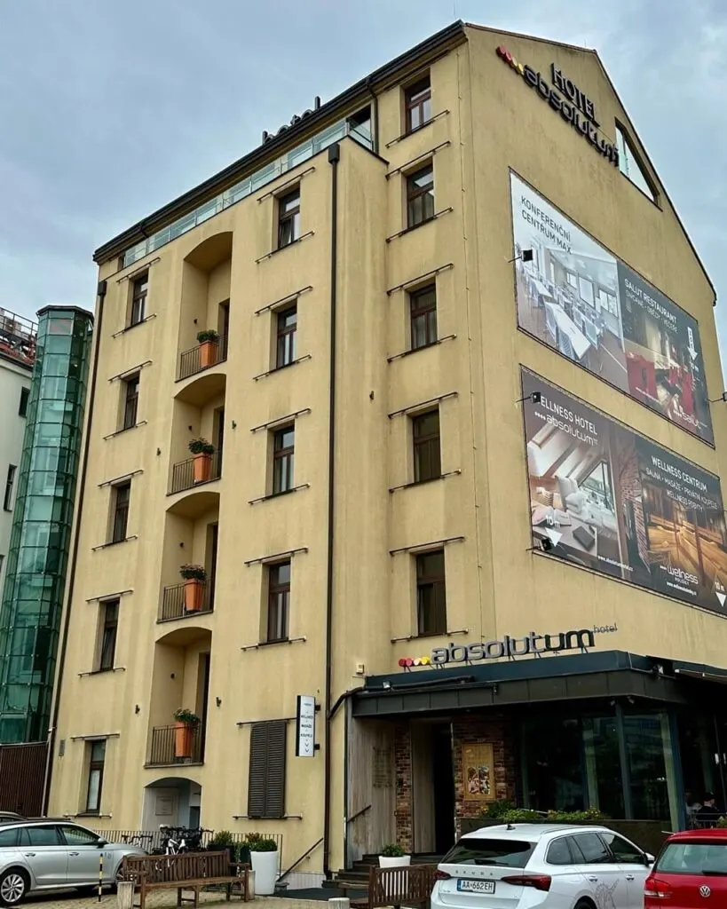 Front view of Absolutum Hotel in Prague with beige exterior, balconies with potted plants, and large advertisements for its amenities.