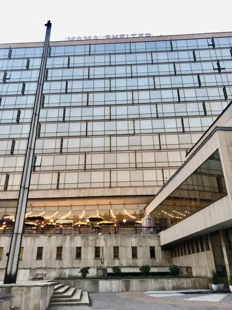 Glass-covered hotel building with “Mama Shelter” signage and outdoor string lights visible through the windows at dusk in Prague.