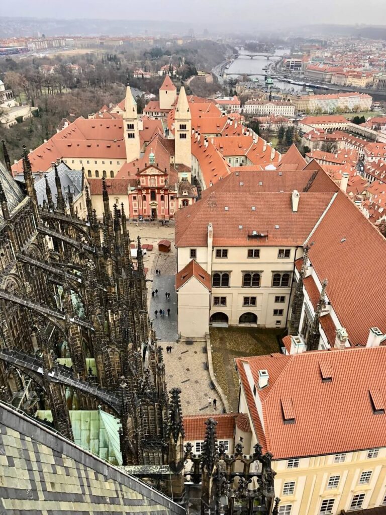 Aerial view from Prague Castle showing medieval rooftops, Romanesque towers, and the Vltava River stretching into the distance.
