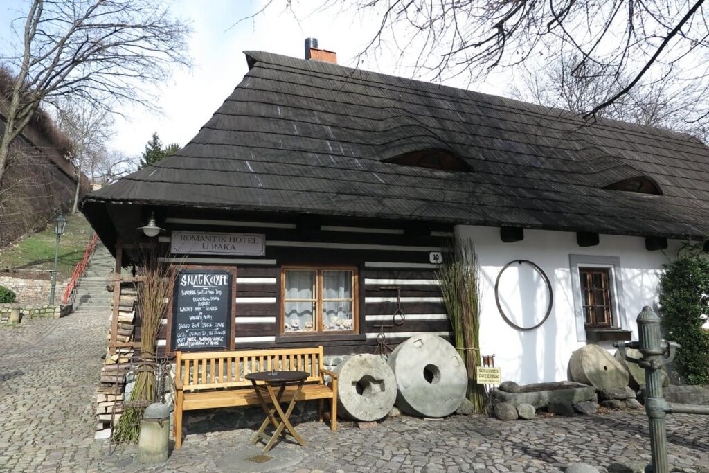 Traditional wooden building with a steep dark roof and stone mill wheels, housing the Romantik Hotel U Raka in Prague.