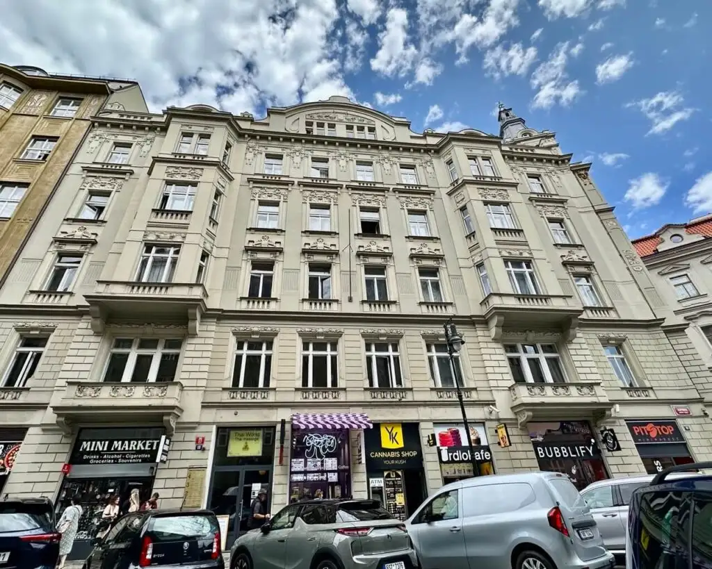 Historic city block in Prague with ornate facades and a mix of businesses at street level including a mini market and cafes.