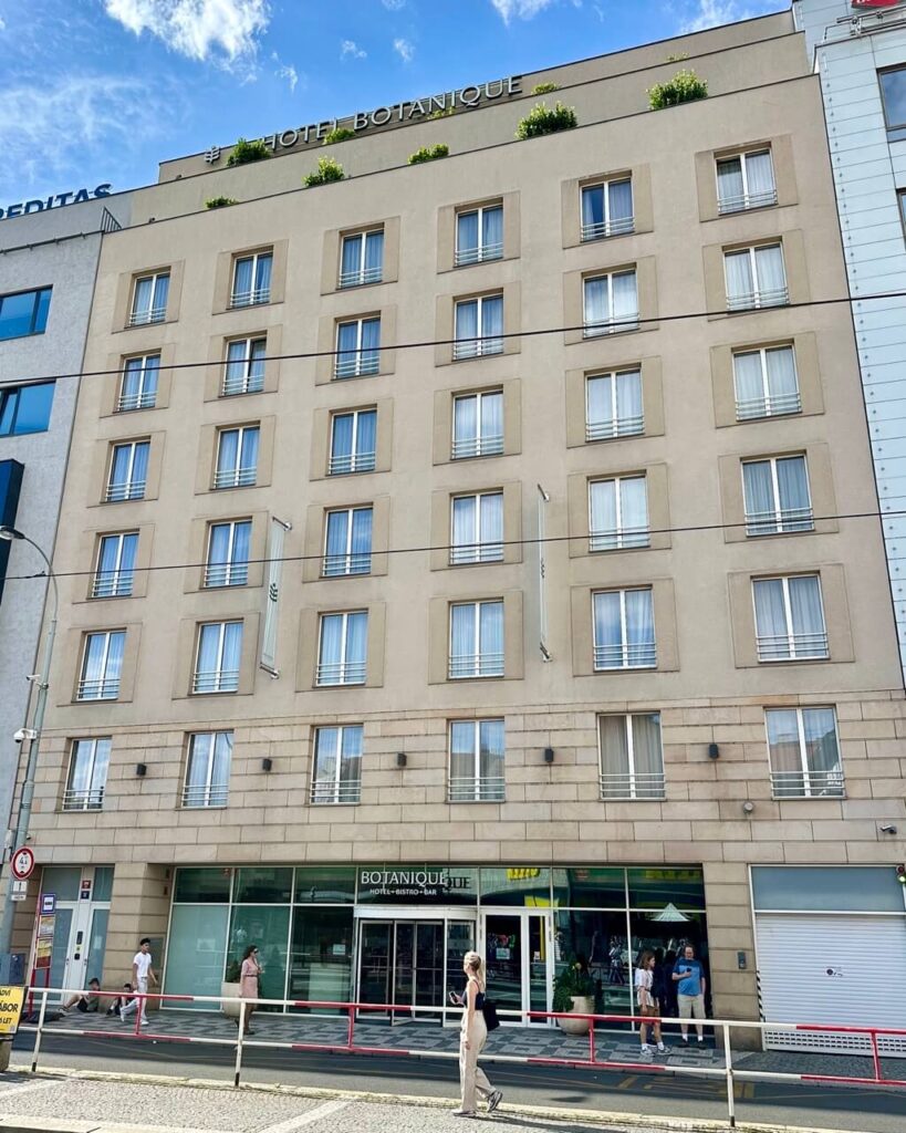 Street view of the beige facade of Hotel Botanique in Prague with its name displayed on the rooftop and a modern glass entrance.