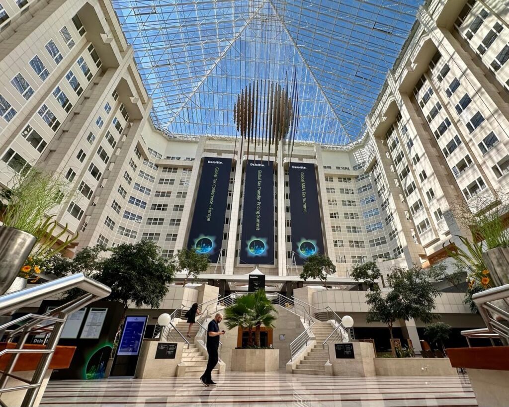 Expansive glass atrium of Hilton Prague with towering ceilings, hanging art installation, and large banners for a Deloitte conference.