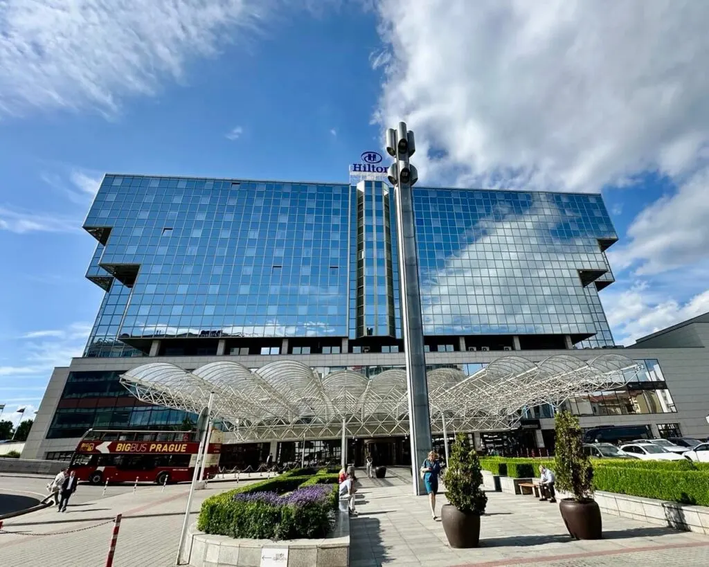 Exterior of Hilton Prague with mirrored glass windows and a futuristic canopy over the entrance, next to a red sightseeing bus.