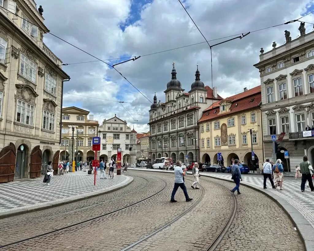 Cobblestone tram-lined street in Malostranské náměstí with baroque architecture, a busy tram stop, and people walking under dramatic skies.