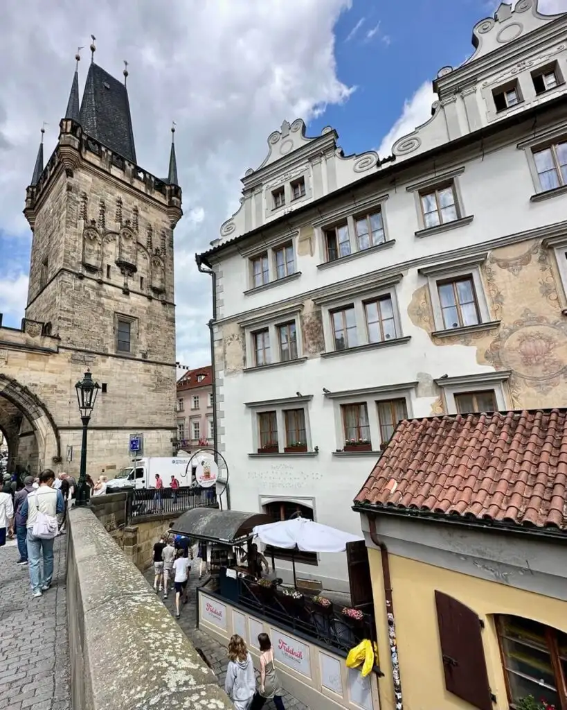View from Charles Bridge showing the Gothic Old Town Bridge Tower and Dům U Tří Pštrosů with its street-level café and frescoed facade.
