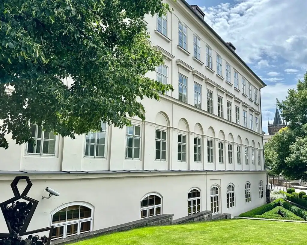 White three-story building with arched windows and manicured lawn, partially hidden by lush green trees in central Prague.