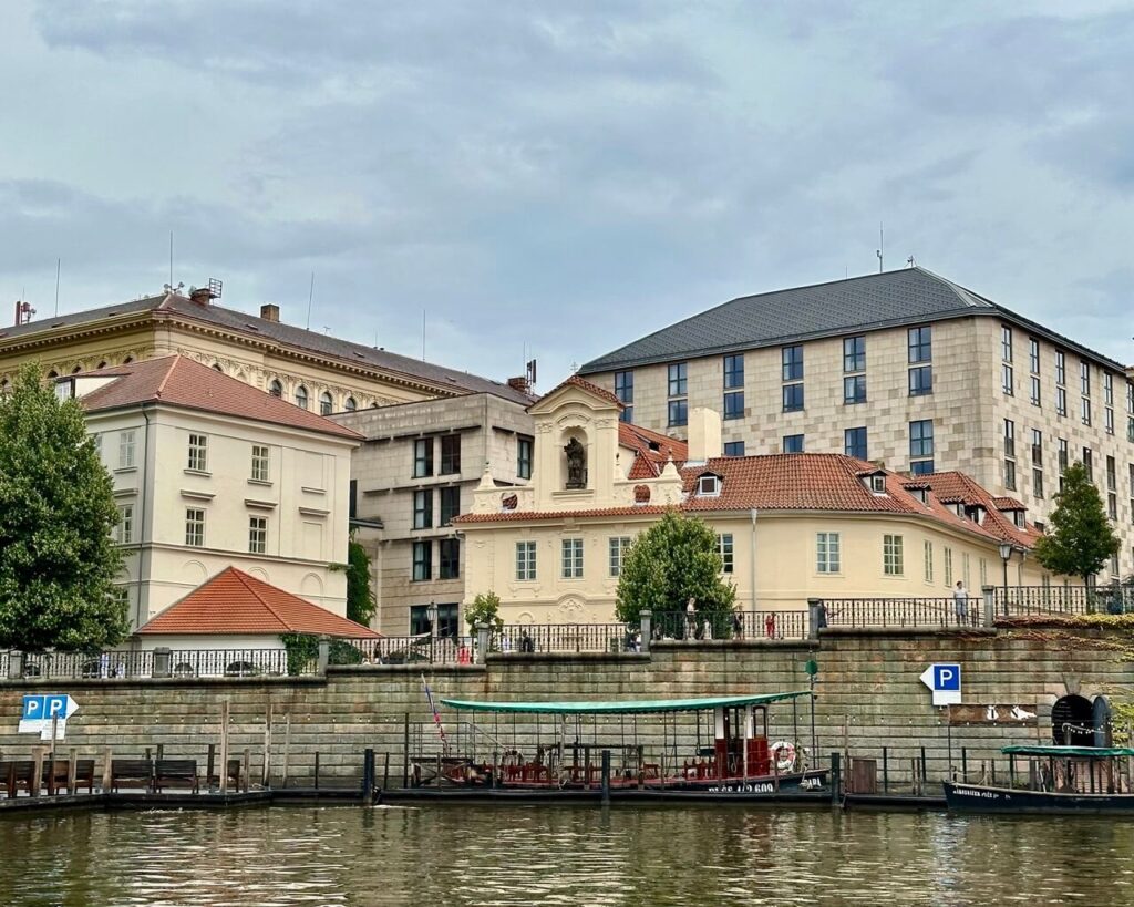 Historic buildings with red tile roofs line the Vltava River across from moored boats and trees under an overcast sky in Prague.