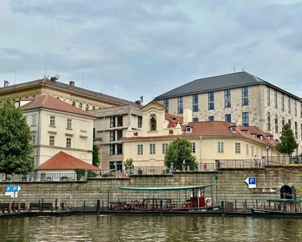 Historic buildings with red tile roofs line the Vltava River across from moored boats and trees under an overcast sky in Prague.