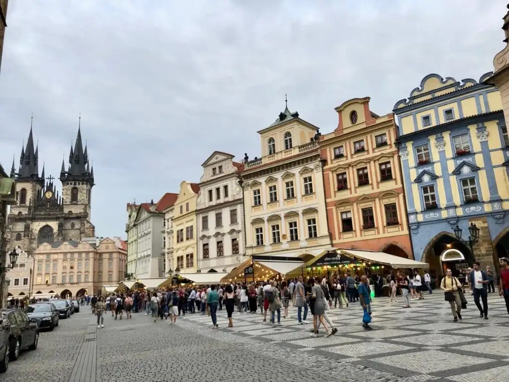 Colorful row of historic buildings in Prague’s Old Town Square with crowds of tourists and the Týn Church in the background.