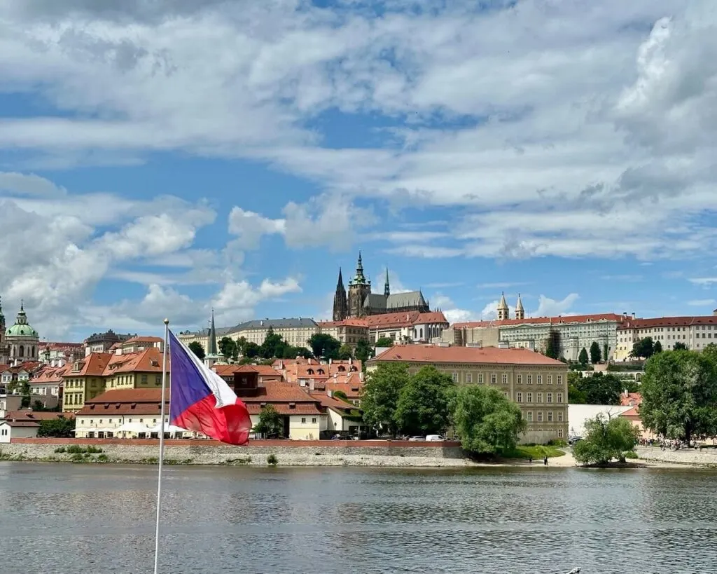 Czech flag waves in the foreground with a panoramic view of Prague Castle and red rooftops across the Vltava River.