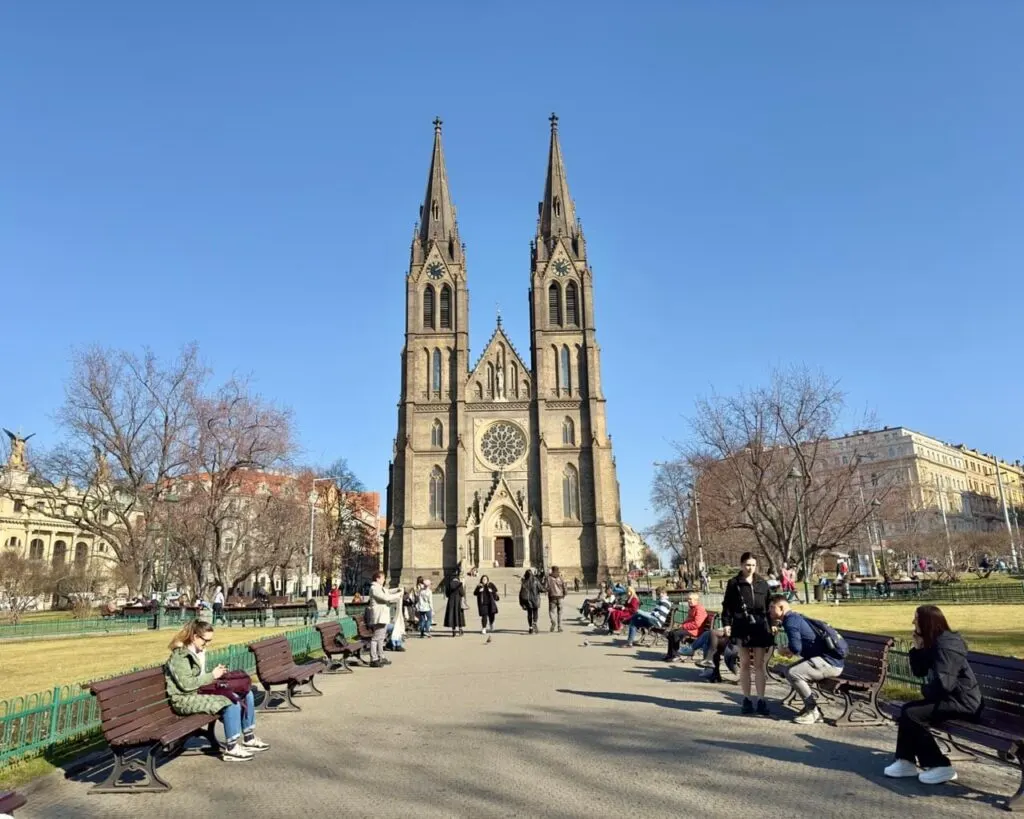 Wide pedestrian path leading to the twin-spired Church of St. Ludmila in Prague, surrounded by benches and people relaxing on a sunny day.
