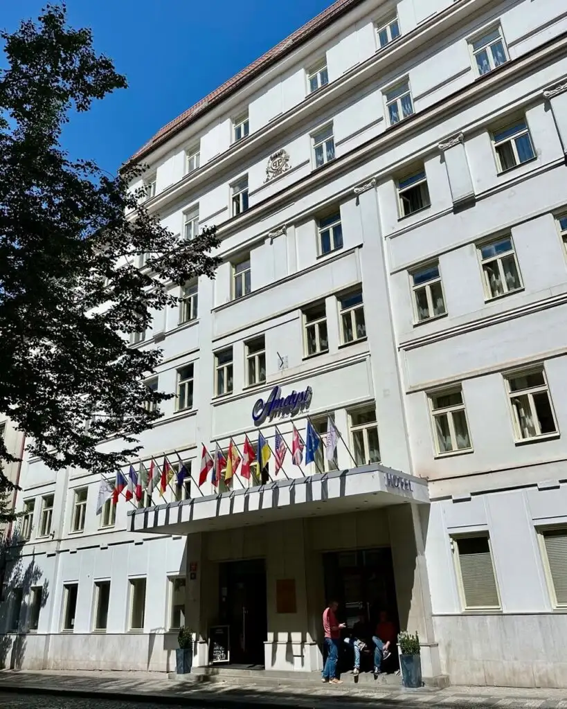 Facade of Hotel Ametyst in Prague, a white mid-rise building with international flags above the entrance and a small group of people outside.