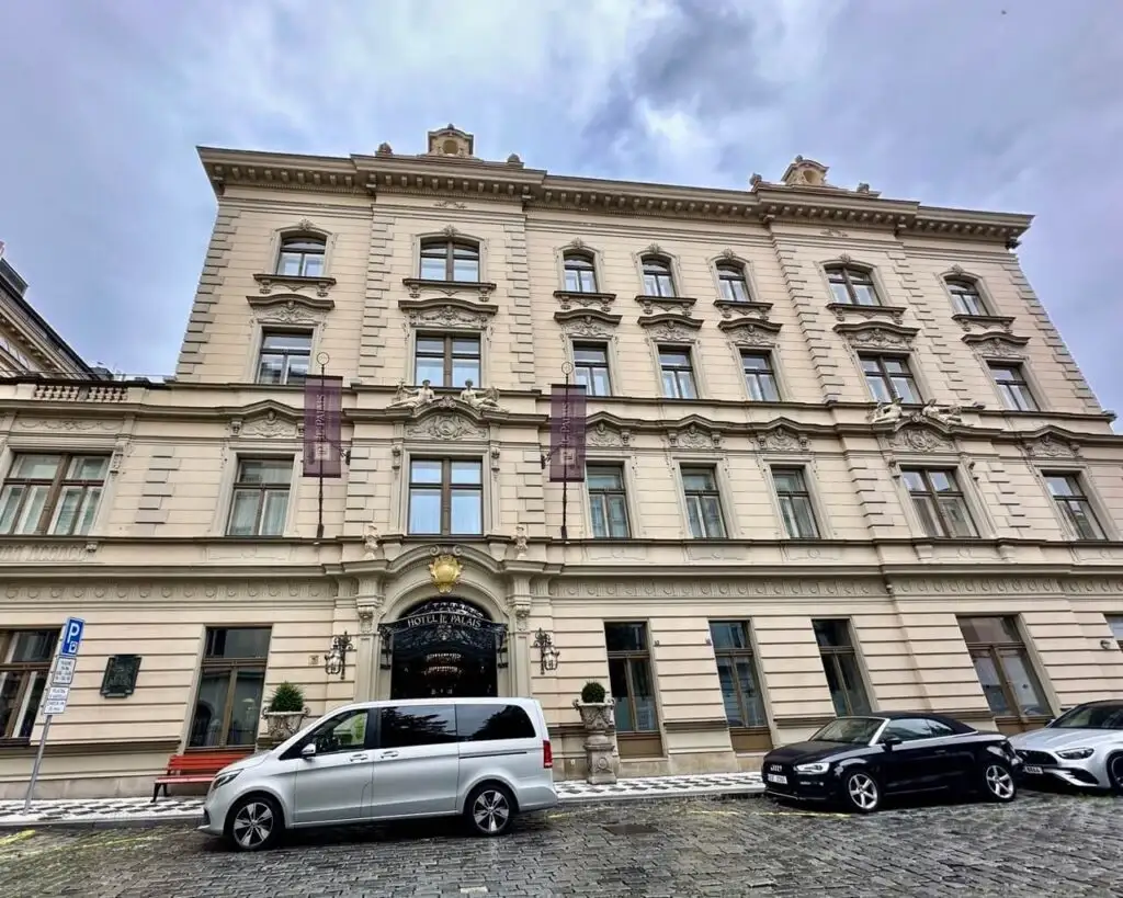Grand historic hotel facade in soft beige tones with detailed molding, arched entry, and luxury cars parked in front.
