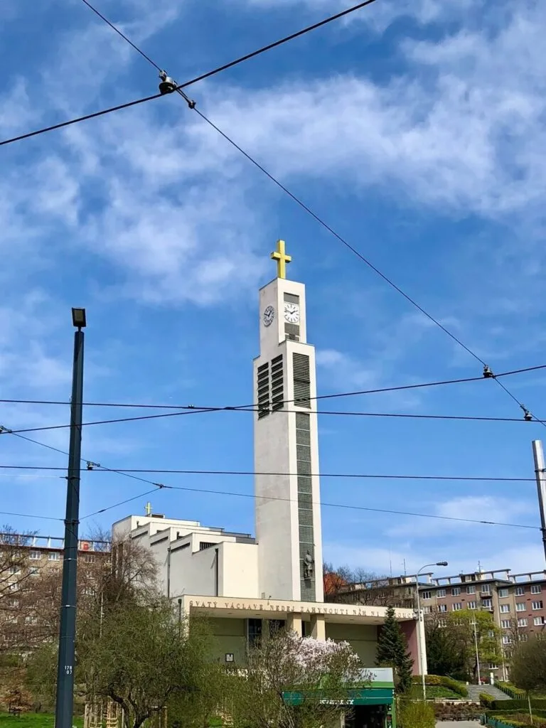 Modern white church tower with two clocks and a gold cross on top, surrounded by power lines and apartment blocks in Prague.