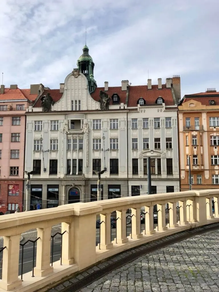Art Nouveau building with copper dome and statues viewed from an elevated stone walkway above a Prague boulevard.