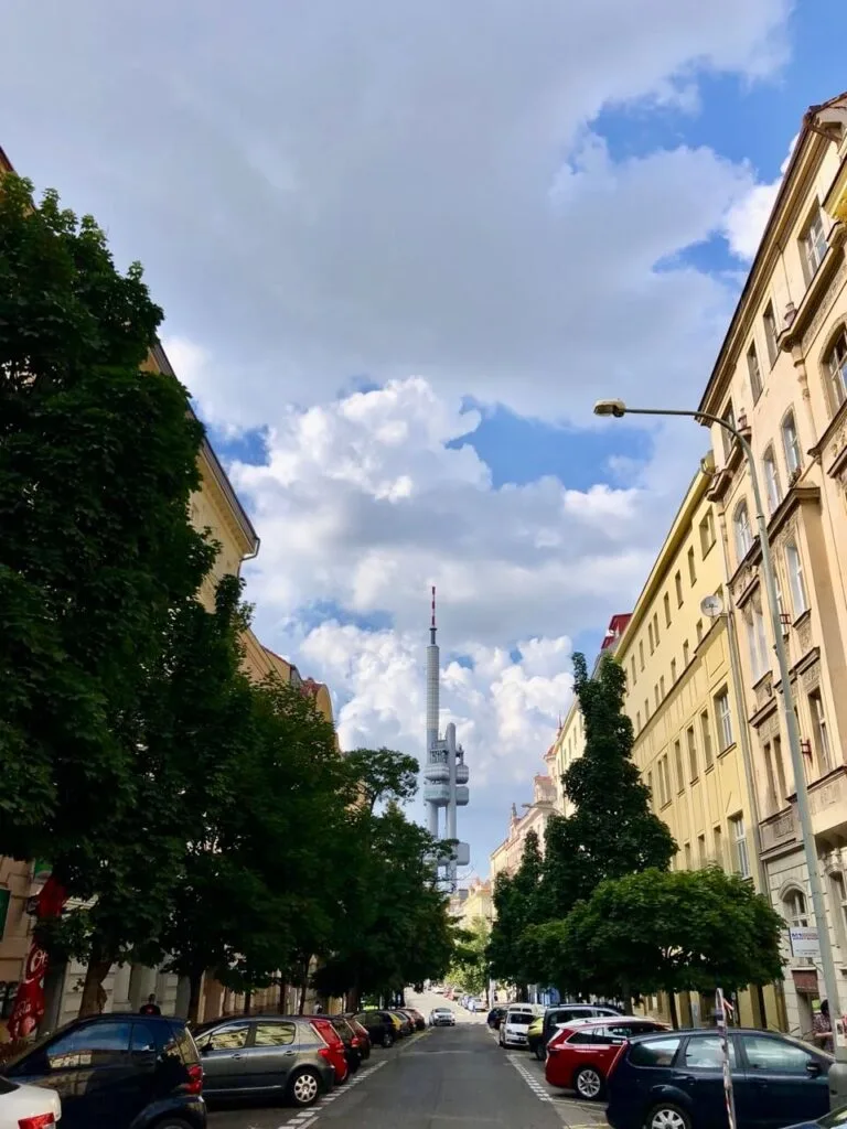 Tree-lined street in Prague with parked cars and a clear view of the futuristic Žižkov TV Tower rising in the background.