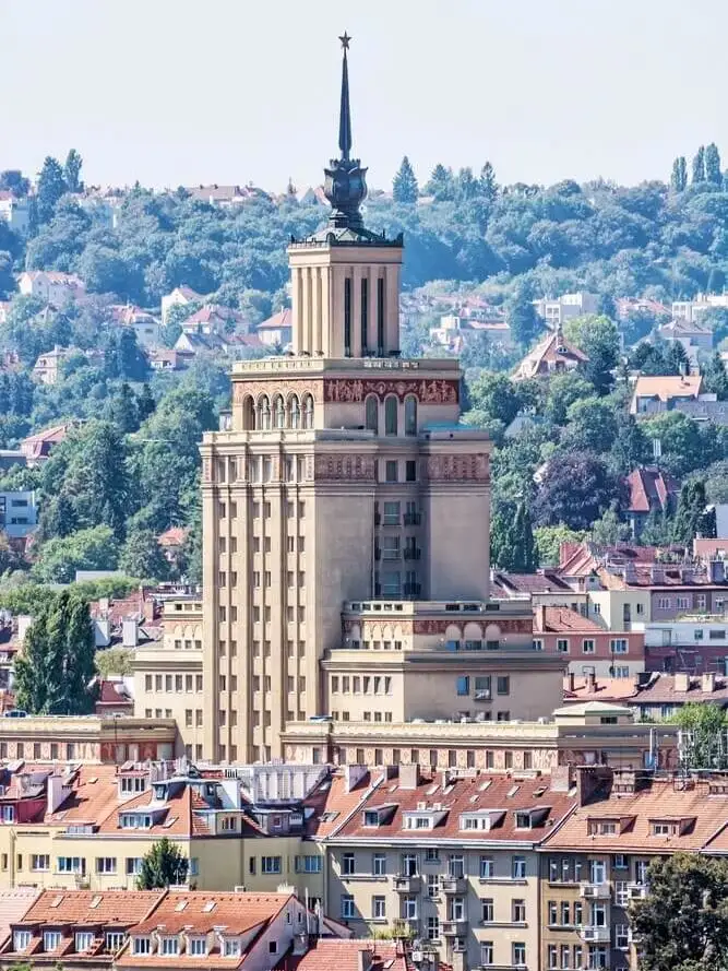Tall, Soviet-era building with a spire and red star, surrounded by red-roofed buildings and greenery in a Prague district.