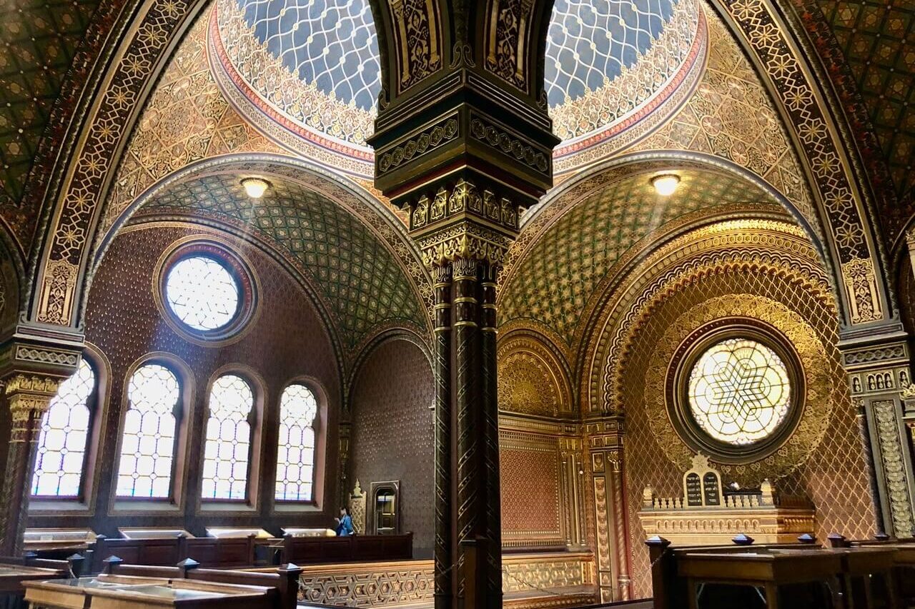 Ornate, gold-accented interior of the Spanish Synagogue in Prague with patterned arches and stained-glass windows.