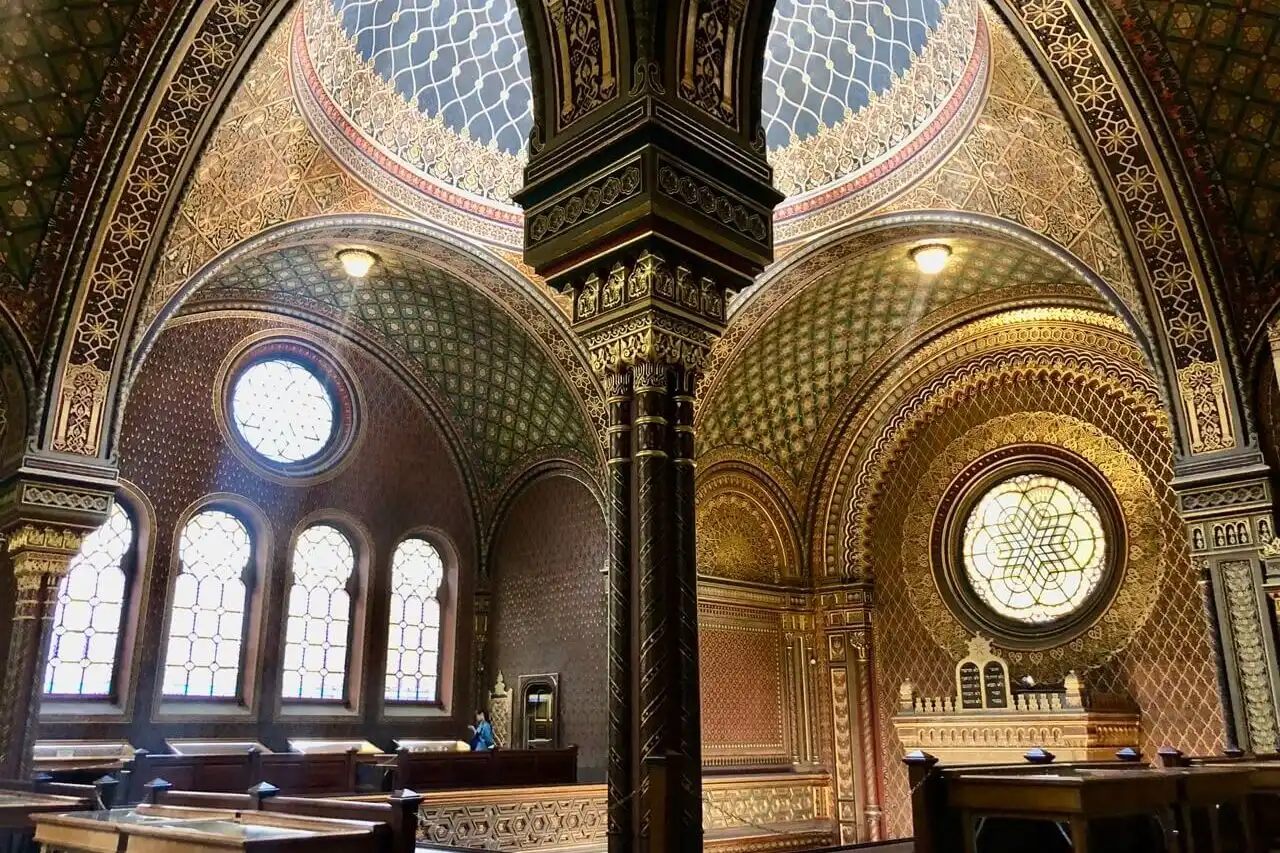 Ornate, gold-accented interior of the Spanish Synagogue in Prague with patterned arches and stained-glass windows.