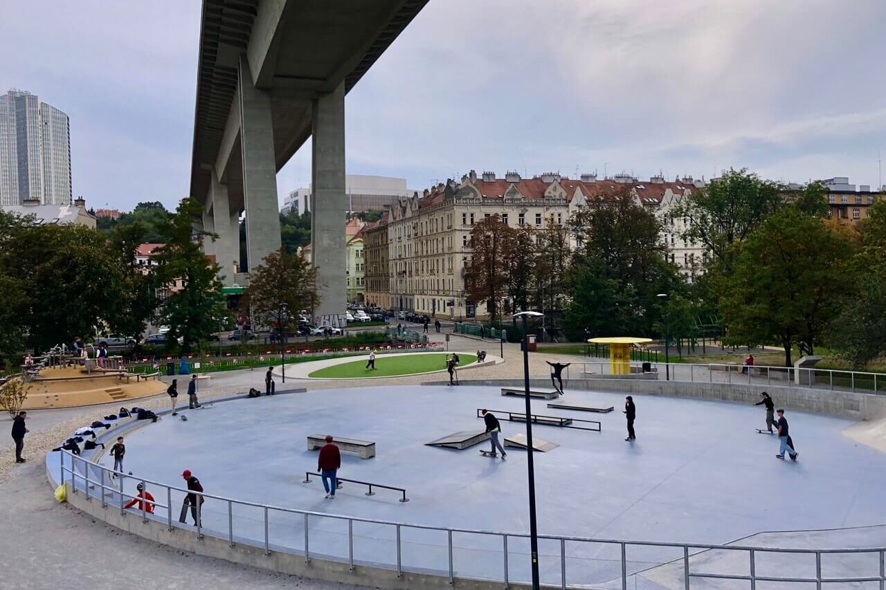 Urban skatepark in Prague beneath the massive concrete spans of Nusle Bridge, with skaters and nearby classic buildings.