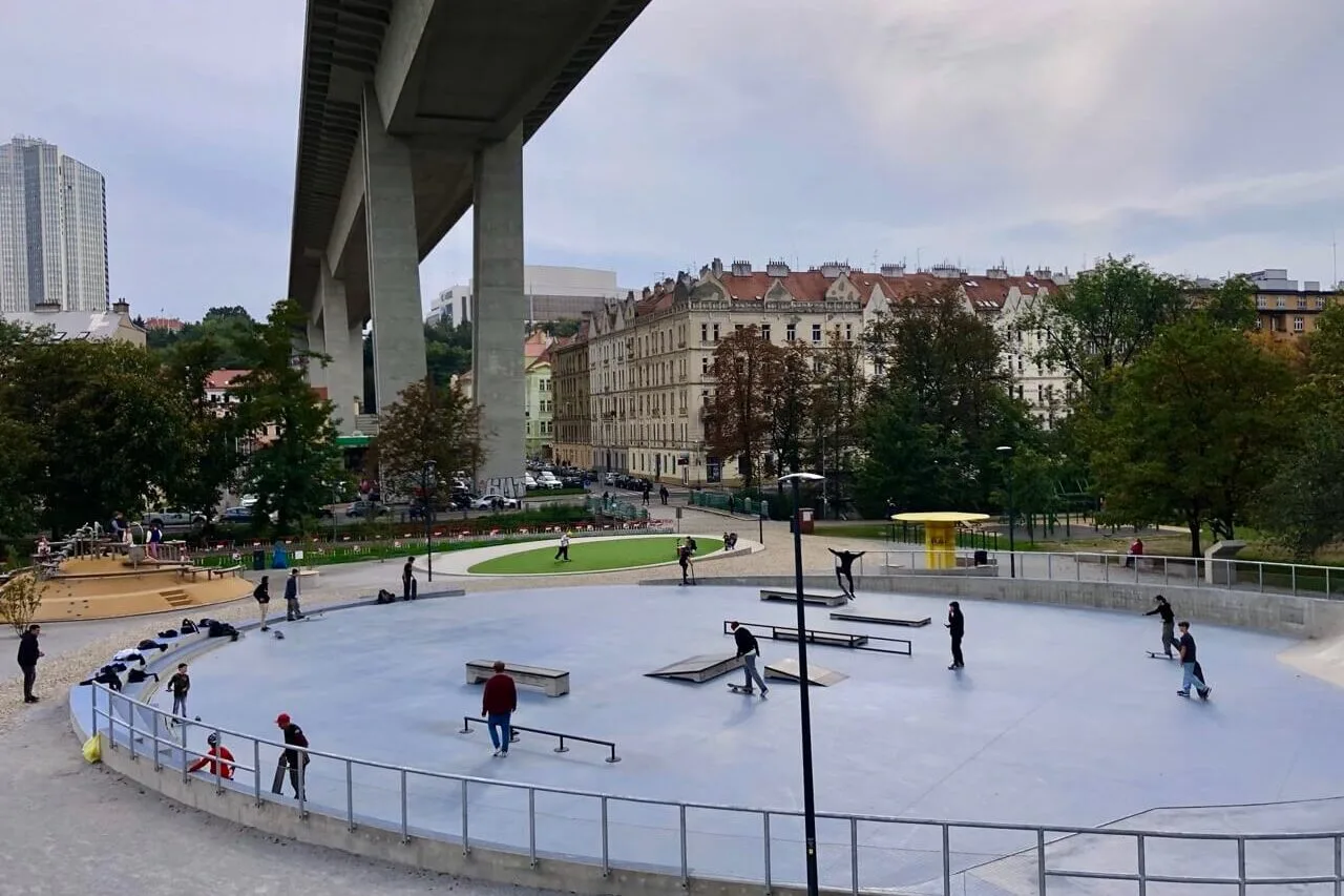 Urban skatepark in Prague beneath the massive concrete spans of Nusle Bridge, with skaters and nearby classic buildings.