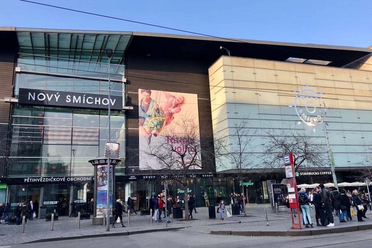 Front entrance of Nový Smíchov shopping mall in Prague with glass facade, large billboard, and tram stop out front.