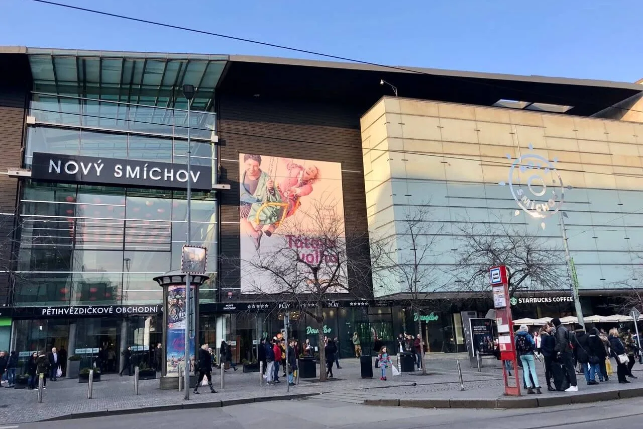 Front entrance of Nový Smíchov shopping mall in Prague with glass facade, large billboard, and tram stop out front.