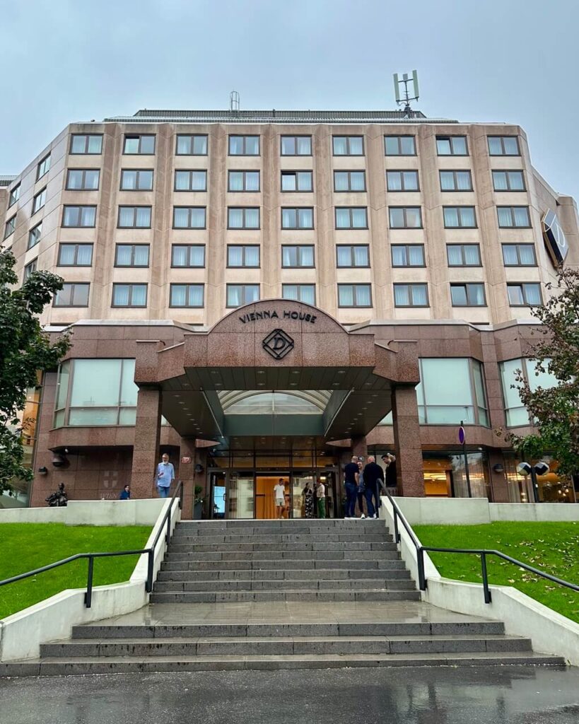 Wide view of Vienna House hotel in Prague with a modern beige facade, symmetrical rows of windows, and a stepped entrance on a rainy day.