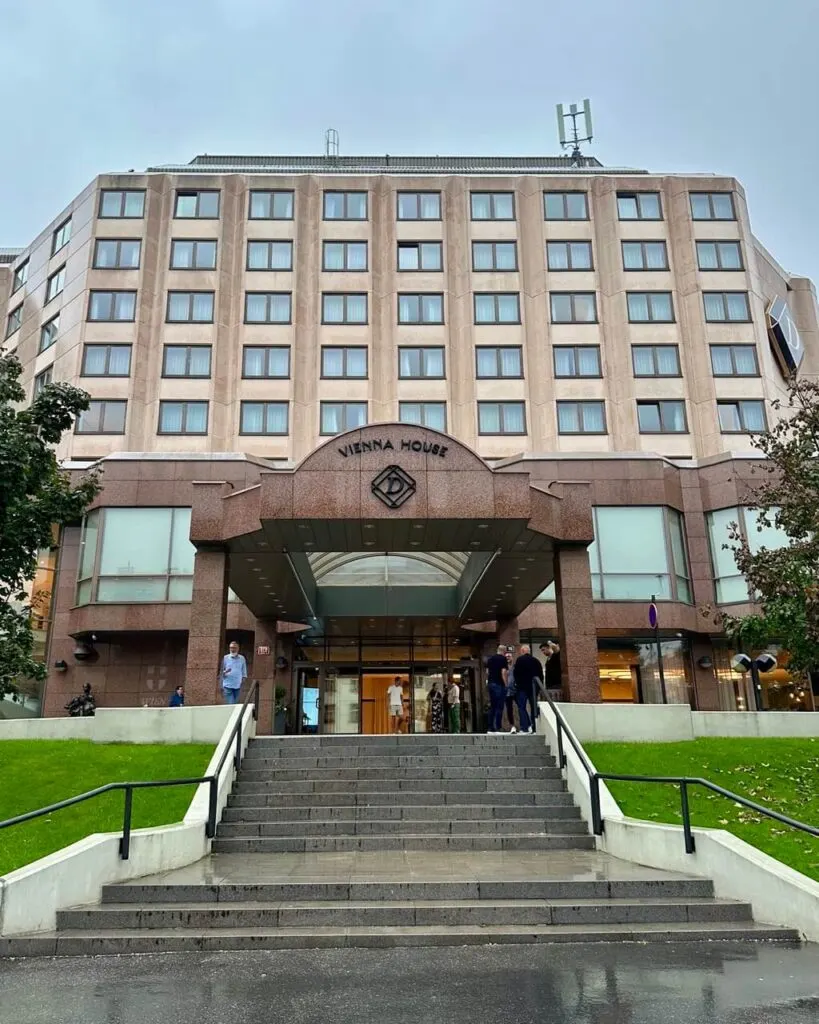 Wide view of Vienna House hotel in Prague with a modern beige facade, symmetrical rows of windows, and a stepped entrance on a rainy day.