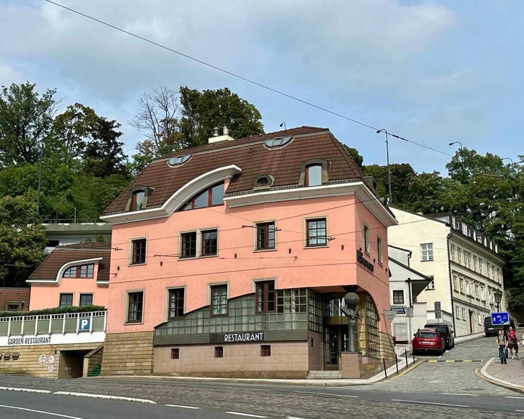 Pink corner building with rounded roof windows and restaurant signage, located on a cobblestone street in a hilly part of Prague.