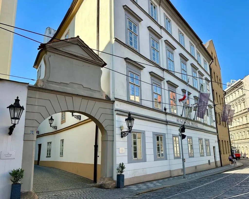 Historic white facade of the Augustine Hotel in Prague with a stone archway, tram wires above, and flags lining the building.