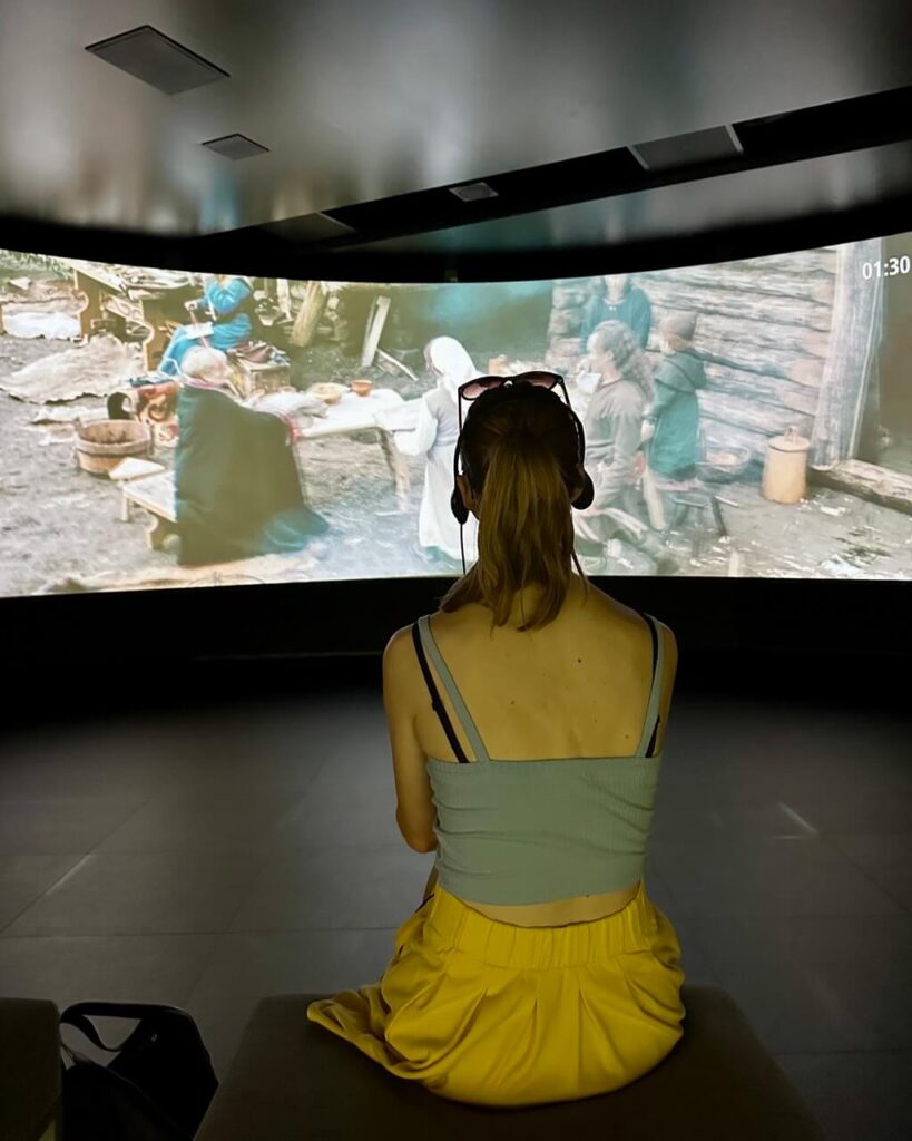A woman in a blue top and yellow skirt watches a historical film inside a dark exhibition room with a curved screen.