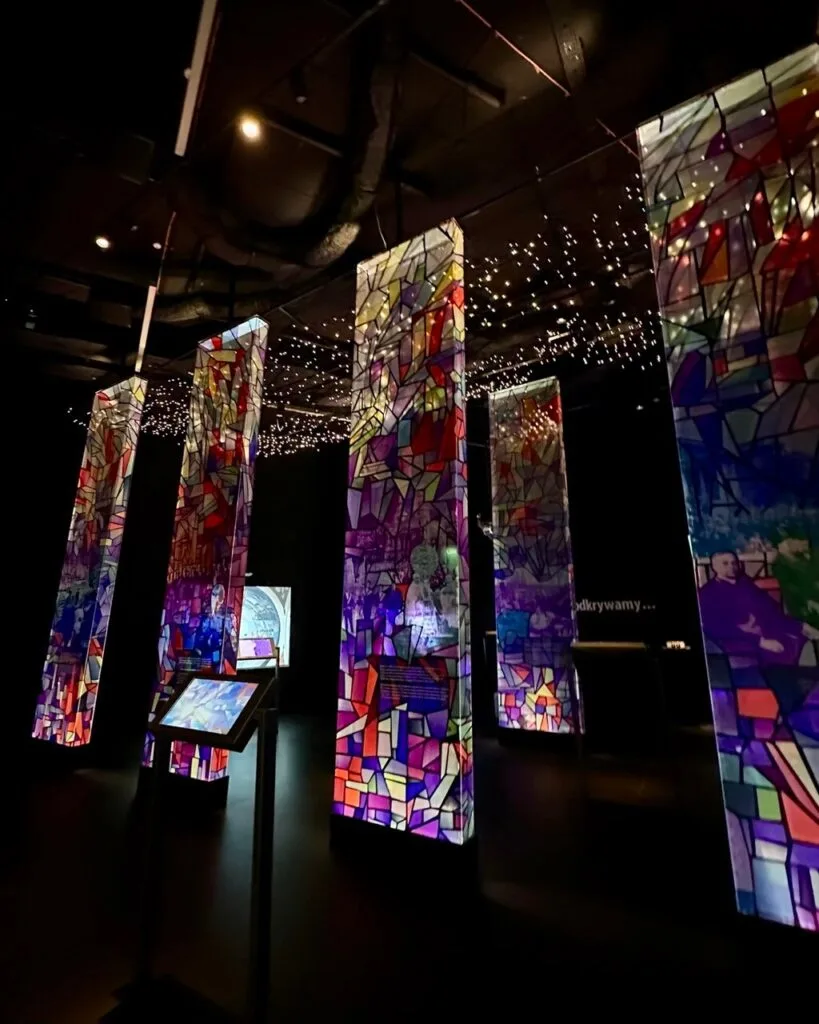 Tall, illuminated columns resembling stained glass art stand in a dark room with star-like lights on the ceiling.