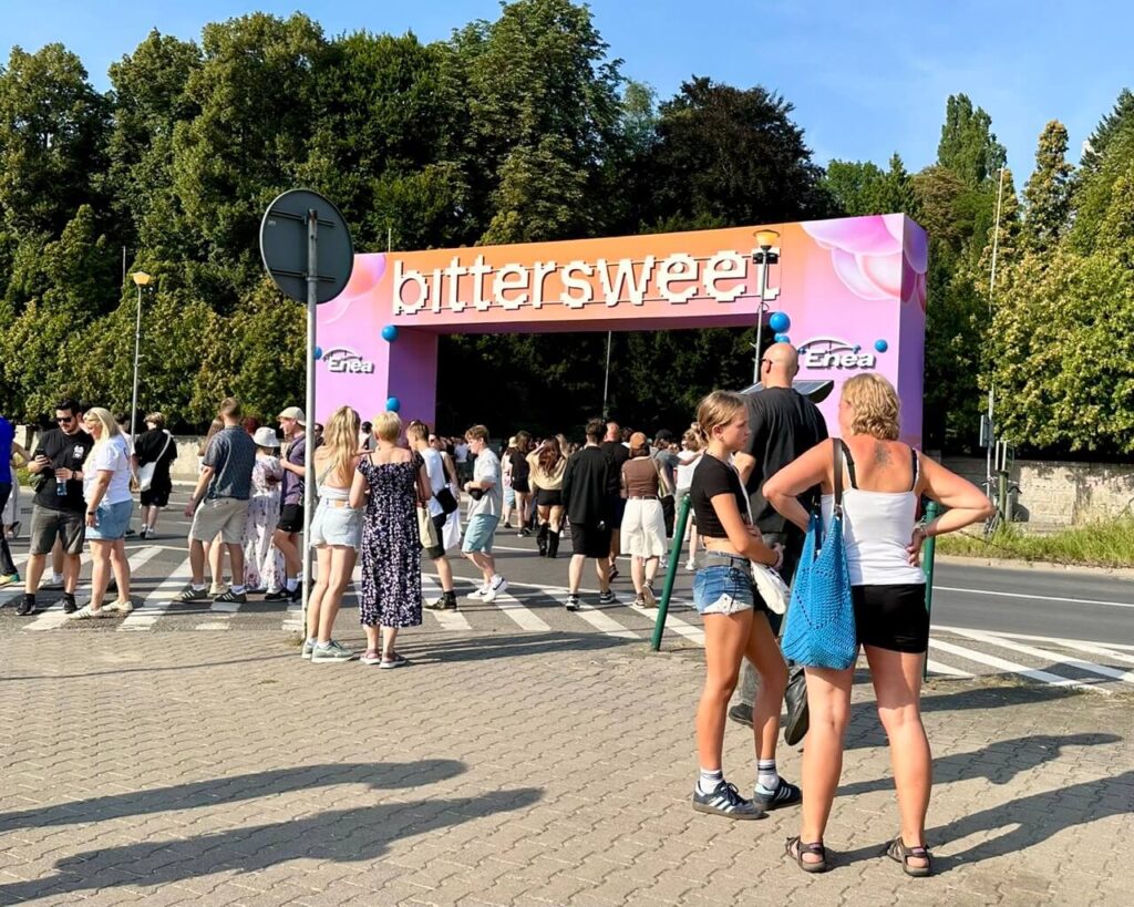 People gather outside a festival gate that reads “bittersweet” on a sunny day.