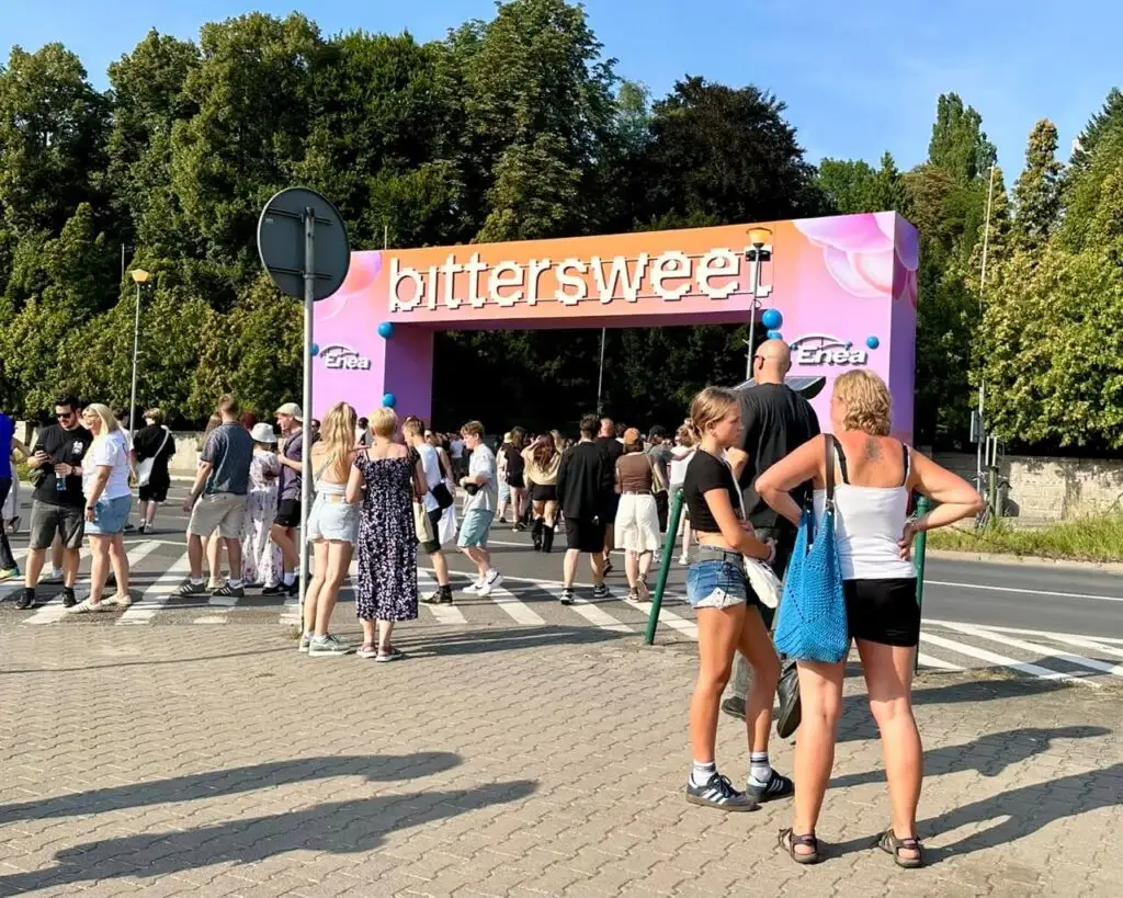 People gather outside a festival gate that reads “bittersweet” on a sunny day.