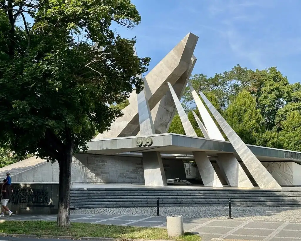 A sharp-angled modern monument made of concrete stands in a plaza, partially shaded by trees.