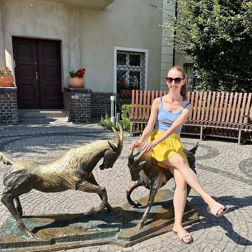 A smiling woman poses while sitting on a bronze goat statue in a sunny courtyard.