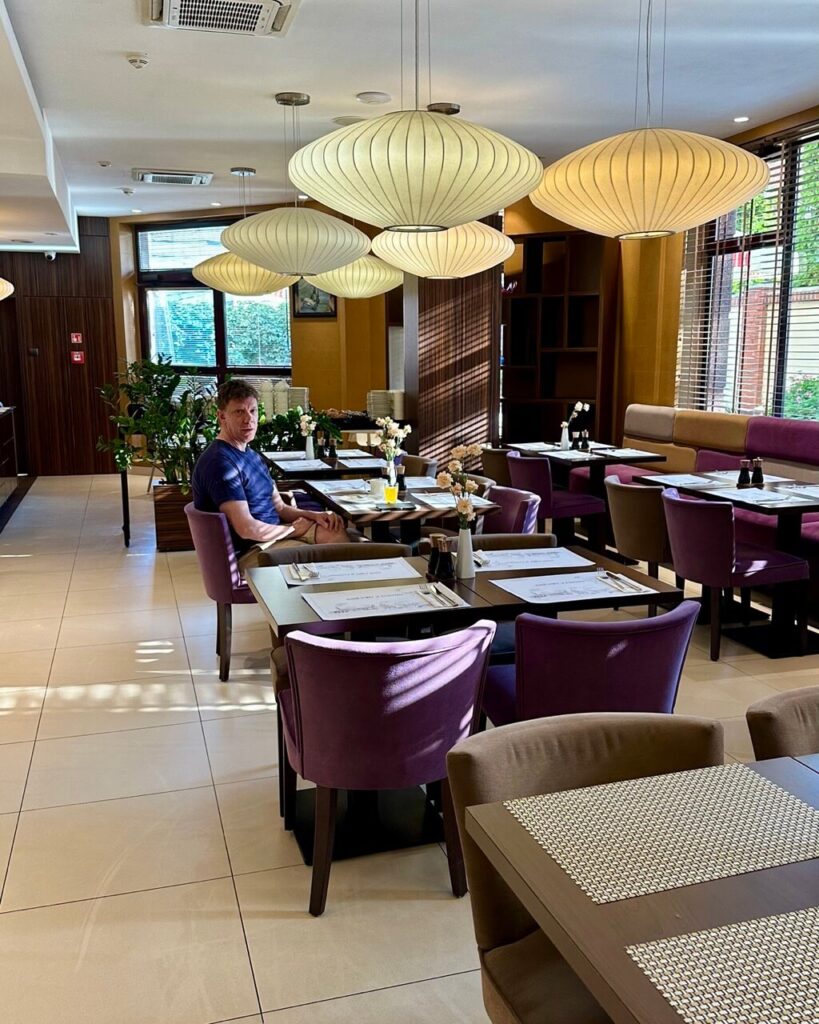 Man sitting alone at a table in a restaurant with modern hanging lanterns and purple upholstered chairs.
