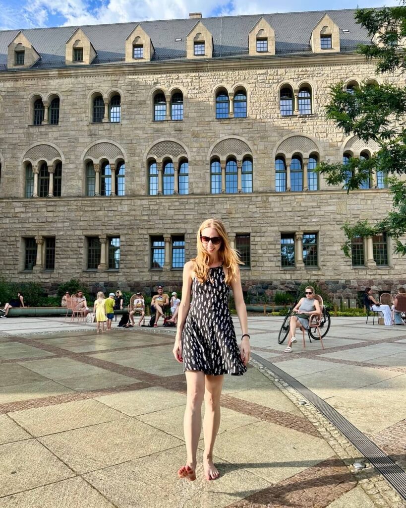 Woman in a sleeveless black dress walking barefoot across a public square in front of a stone building.
