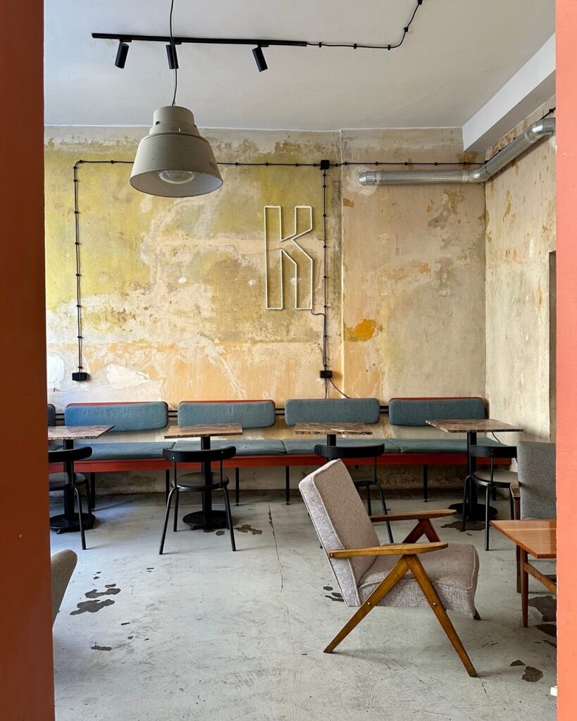 Minimalist interior of a hip café with mismatched furniture and exposed plaster walls featuring a neon “K.”