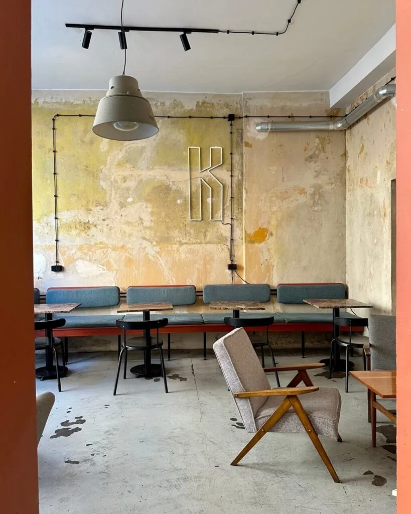 Minimalist interior of a hip café with mismatched furniture and exposed plaster walls featuring a neon “K.”