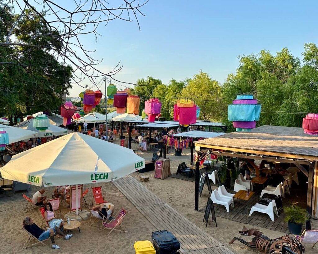 Outdoor beach-style bar with umbrellas, colorful lanterns, and people relaxing on sand under the evening sun.