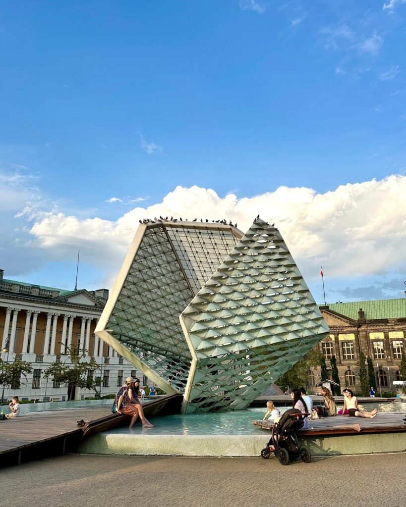 People relax around a modern geometric fountain with pigeons perched on top in a public square.